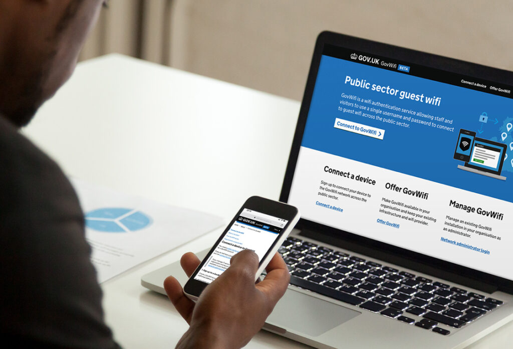 Photo showing a man viewing the GovWifi BETA website on a laptop and a mobile phone