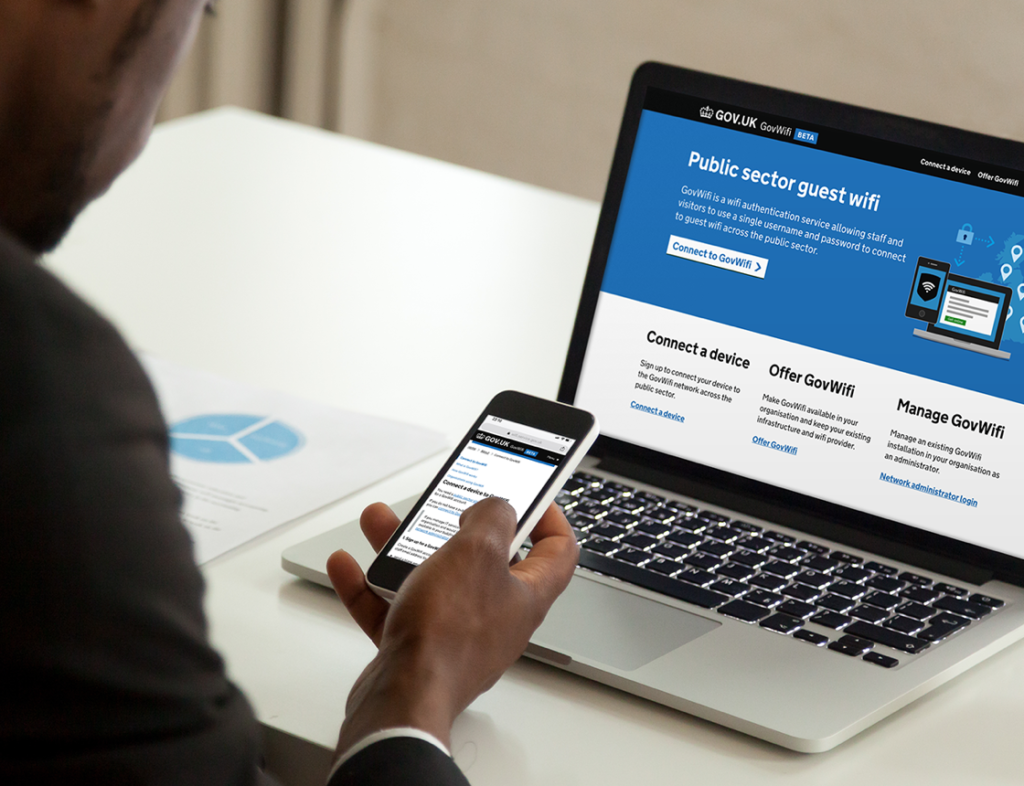 Photo showing a man viewing the GovWifi BETA website on a laptop and a mobile photo