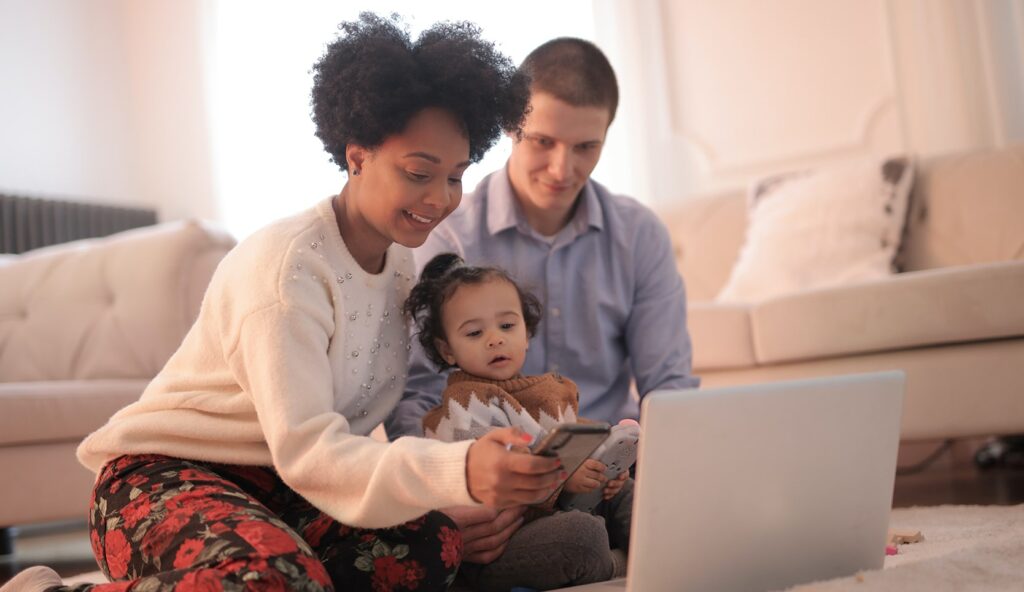 Image of woman, man and a baby sitting on the floor of a living room in front of a laptop and phone