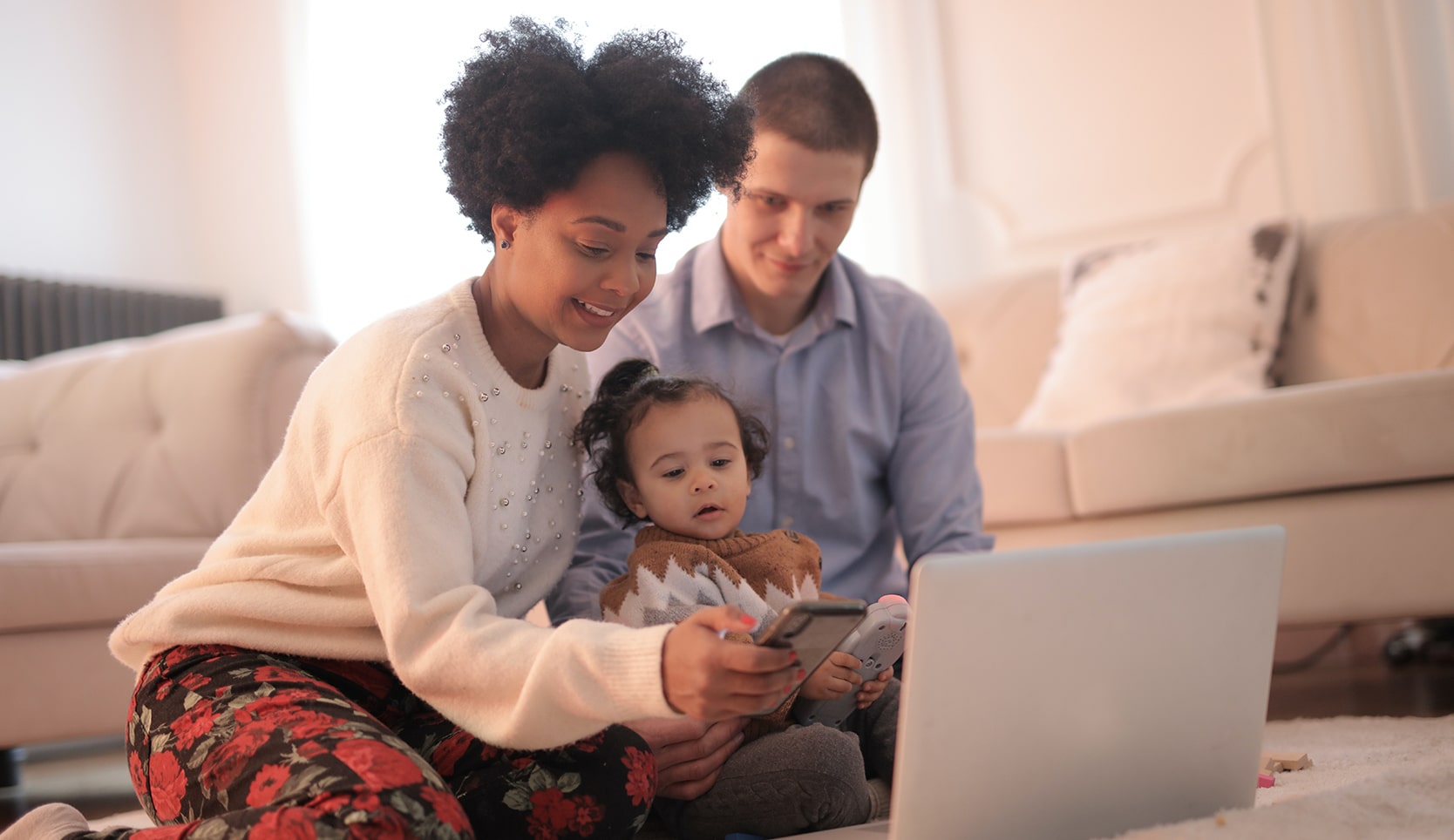 Image of woman, man and a baby sitting on the floor of a living room in front of a laptop and phone