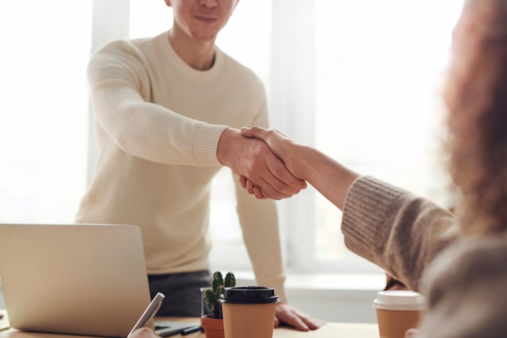 A man and a woman shake hands across a table with a laptop and coffees