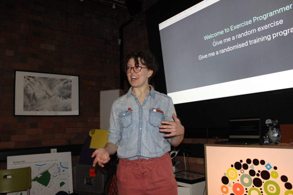 The post author Evie Skinner presenting in front of a display screen at a Queer Tech Bristol meeting. She is wearing a blue denim shirt and red trousers.