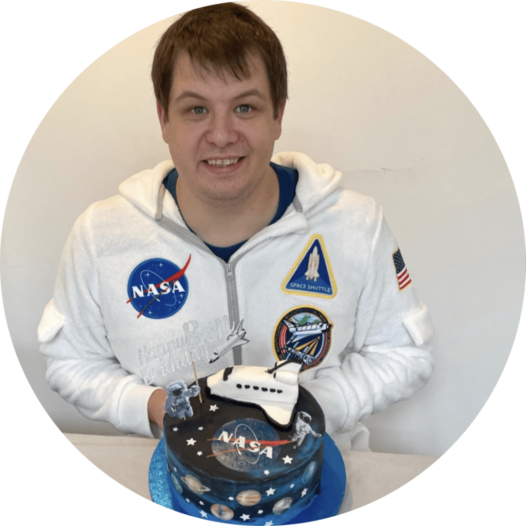 Image of Ben Noble wearing a NASA suit behind a NASA themed cake