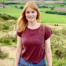 Helena is standing outside in a green field, she is wearing a red top and had long red hair. She is smiling at the camera.