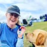 Nicola smiles at the camera, she is wearing a blue cap and blue top, she is next to a golden labrador.