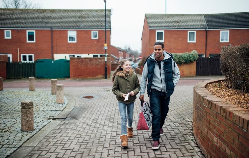 A man and a young girl walking through a residential neighborhood with red-brick houses, both smiling and holding shopping bags on a cloudy day.