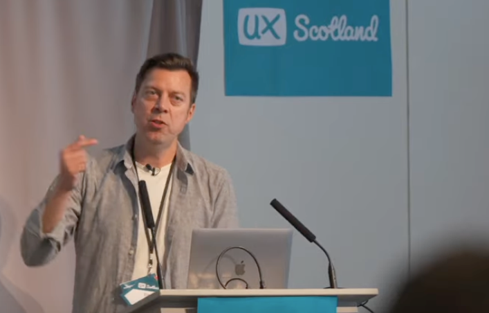 Dan Healy stands in front of a UX Scotland sign, he is delivering a talk and speaking into a microphone