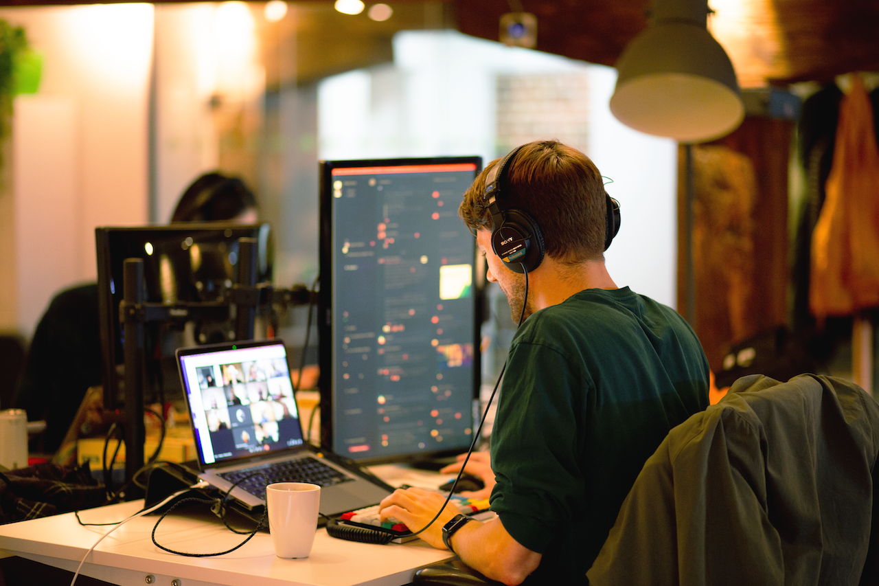 Man sitting at a desk in front of a laptop and monitor, wearing headphones while joining a virtual teams call.